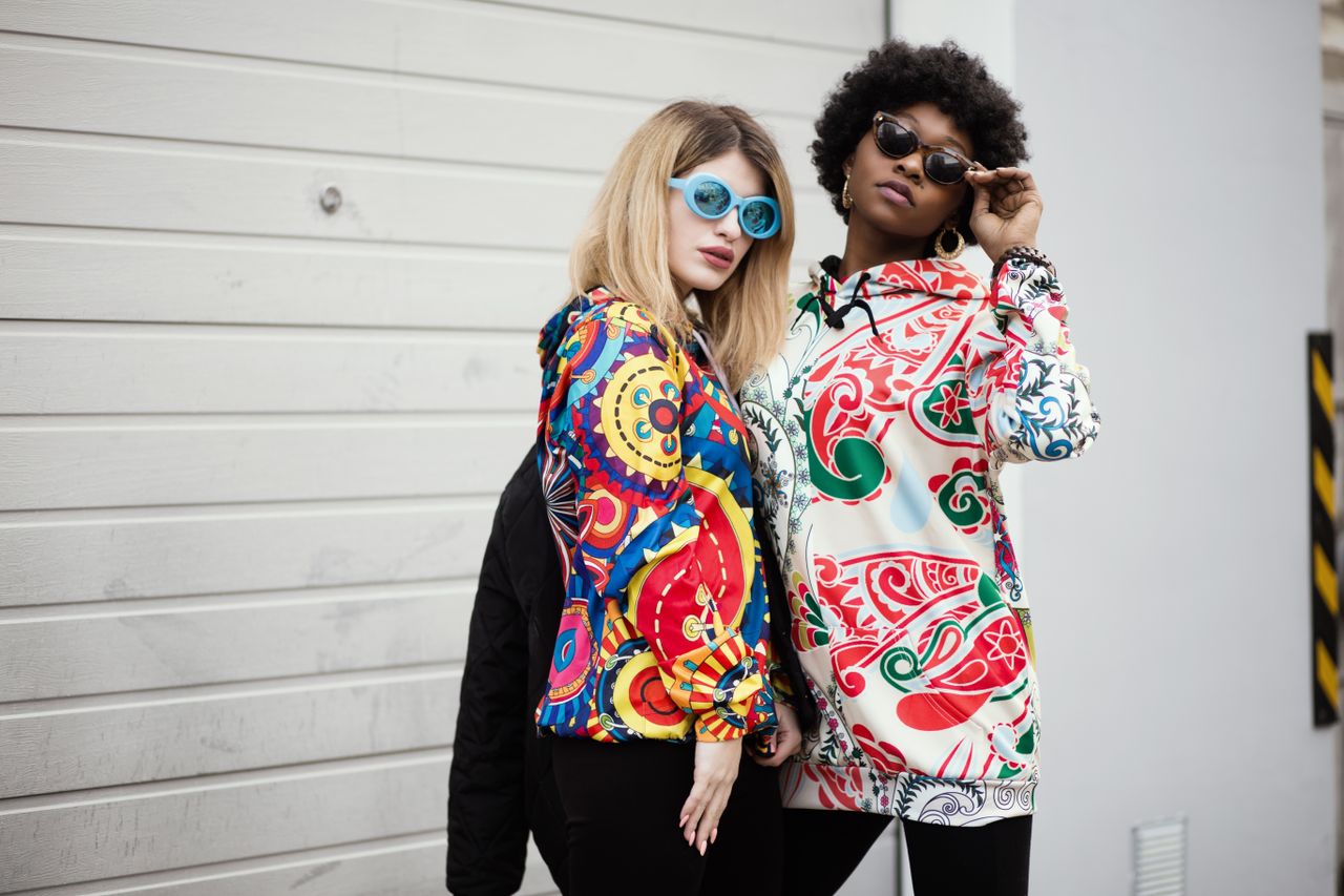 gallery-05 Two stylish women wearing colorful graphic hoodies and sunglasses against a modern urban backdrop.