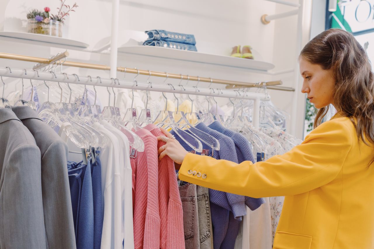 gallery-02 Woman browsing clothes on a rack in a clothing store, wearing a stylish yellow blazer.