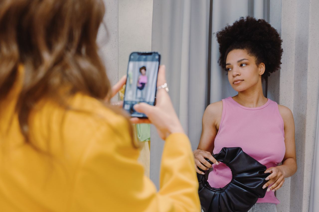 gallery-03 Woman in pink top poses with handbag while another takes a smartphone photo in a changing room.