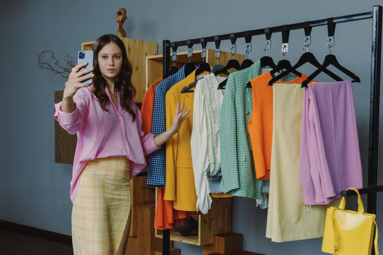 gallery-01 Woman in colorful outfit taking a selfie amidst vibrant clothing options in a modern boutique.